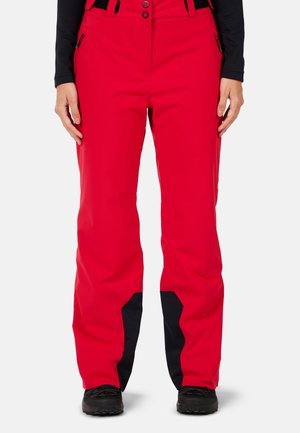 Person trägt leuchtend rote Schneehose mit schwarzen verstärkten Patches an den Knöcheln, schwarze Stiefel und ein schwarzes langärmeliges Oberteil.