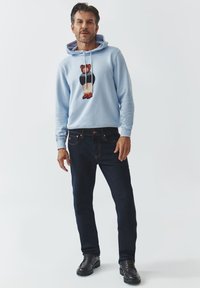 Sudadera azul clara con gráfico de un oso, capucha con cordones, combinada con jeans azul oscuro y zapatos negros. Textura suave y ajuste casual.