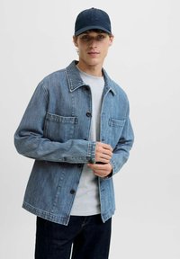Veste en denim bleu avec une finition texturée, fermeture à boutons et deux poches avant, portée sur une chemise gris clair et un jean foncé.