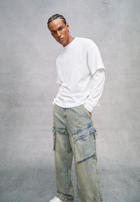Camisa de manga larga blanca combinada con pantalones cargo azul claro con bolsillos. El modelo está de pie con las manos en los bolsillos, frente a un fondo gris.