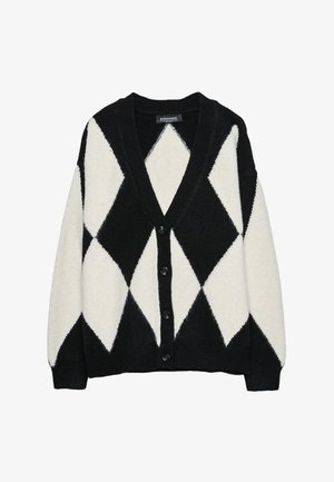 Femme portant un cardigan en tricot à losanges noir et blanc avec un short assorti, des chaussettes noires et des mocassins noirs, debout contre un mur gris.