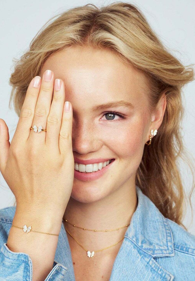 Guldsmycken med fjärilsdesign, inklusive en ring, armband och halsband. Modellen bär en denimjacka och har mjukt, vågigt hår.