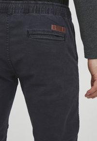 Navy blue cotton blend pants with elastic waistband, rear pocket detail, and a brown leather patch labeled "INDICODE." Soft texture and casual design.