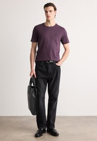 Tricou din bumbac violet cu un mic logo, asociat cu blugi negri din denim. Ține în mână o geantă din piele neagră, poartă pantofi negri și un centură.