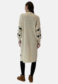 Lång krämfärgad cardigan i grovstickat med svarta hjärtan på ärmarna, i kombination med svarta leggings och tanfärgade ankelboots.
