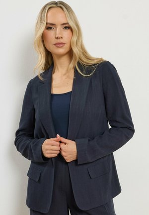 Lyshåret kvinde iført navyblå blazer og top, knapper blazeren med begge hænder, foran en ensartet hvid væg.