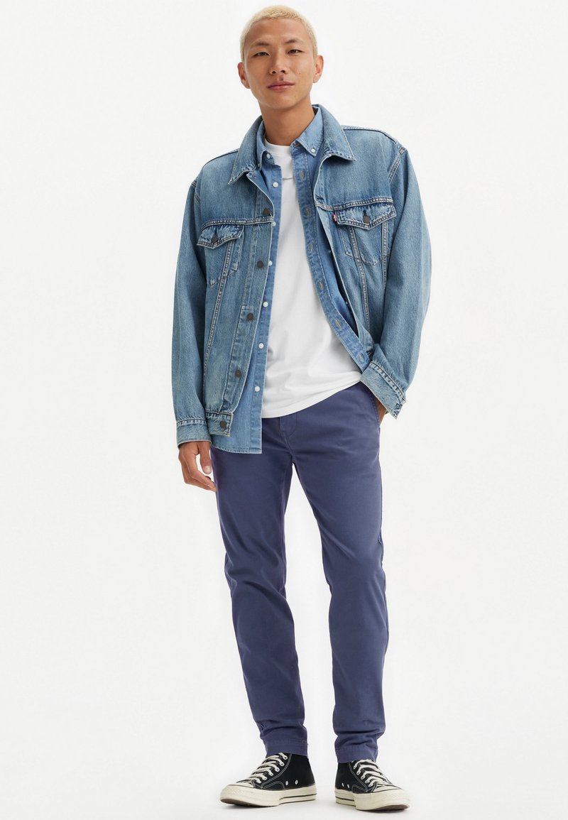 Chaqueta de denim en azul claro, camiseta blanca debajo, pantalones azul marino y zapatillas altas negras con suelas blancas. Atuendo casual, sin patrones.