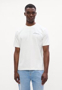 Tricou alb cu mânecă scurtă din bumbac, cu guler rotund și un logo mic imprimat în negru pe pieptul stâng. Purtați cu blugi de culoare albastru deschis.