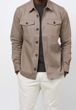 Homme portant une chemise taupe boutonnée sur un t-shirt noir avec un pantalon blanc, mains dans les poches, devant un fond clair uni.