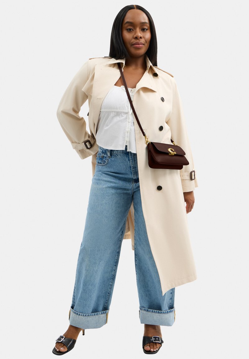 Femme portant un trench-coat beige, un chemisier blanc, un jean large avec revers, des talons ouverts noirs et un sac bandoulière marron foncé, debout avec assurance.