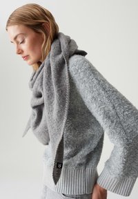 Maglione oversize grigio chiaro con finitura texturizzata e polsini a costine, abbinato a una morbida sciarpa grigia drappeggiata attorno al collo.