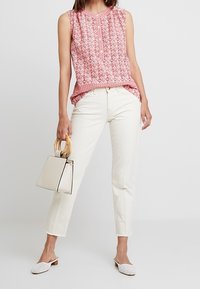 Top rosso senza maniche con motivo, pantaloni bianchi a gamba dritta, mule bianchi intrecciati e una piccola borsa beige con manico in legno.