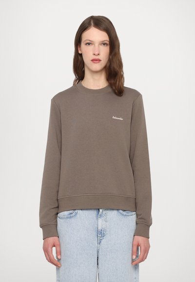 Sweatshirt marron avec col rond, poignets et ourlet côtelés, affichant un petit logo brodé sur la poitrine. Associé à un jean bleu clair.