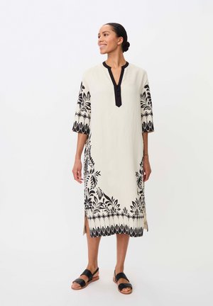 Vrouw staat in een witte midi-jurk met zwarte bloemenpatronen op mouwen en zoom, gecombineerd met zwarte sandalen, glimlachend en naar rechts kijkend.