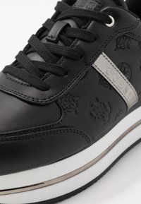 Czarne skórzane trampki z wytłoczonymi wzorami logo, srebrnym paskiem akcentującym i czarnymi sznurowadłami. Charakteryzują się białą podeszwą z metalicznymi detalami.