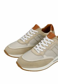 Paire de baskets beige et crème avec des lacets blancs, des accents en cuir marron au talon et à la languette, et des semelles en caoutchouc.