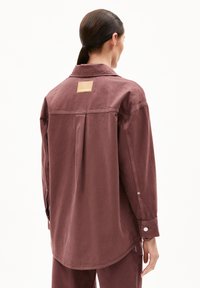 Frau mit dunkel zurückgebundenem Haar, die ein übergroßes mauvefarbenes Hemd mit Knöpfen und passende Hosen trägt, von hinten auf weißem Hintergrund gezeigt.