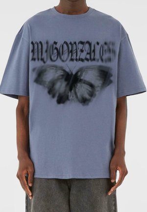Person trägt ein locker sitzendes blaues T-Shirt mit schwarzem gotischem Text und einer verschwommenen schwarzen Schmetterlingsgrafik auf der Vorderseite.