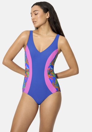 Mujer con cabello largo y oscuro, vestida con un traje de baño de una pieza azul con paneles rosas y patrones florales de colores en los lados, de pie con las manos detrás de la espalda.