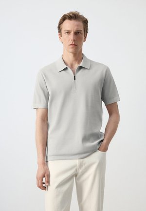 Homme portant un polo gris clair à manches courtes avec fermeture éclair sur le devant et un pantalon écru, debout devant un fond blanc uni.