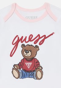 Weißer Baumwoll-Body mit einem Cartoon-Bären, der einen roten Pullover mit einem dreieckigen Logo trägt. Heller, rosa eingefasster Kragen. Roter "guess"-Text darüber.