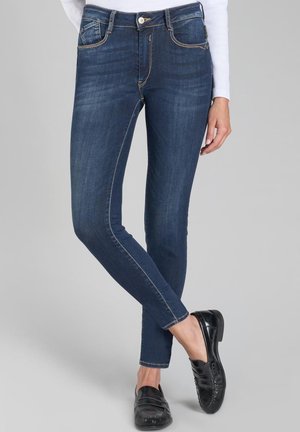 Jeans blu scuro aderenti in tessuto denim con tasche frontali, passanti per cintura e dettagli di cucitura. Abbinati a scarpe nere, piane e senza lacci.