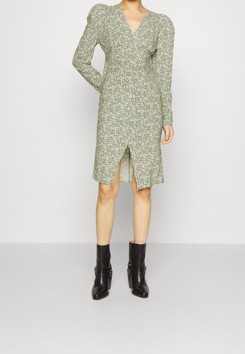 Grünes Blumenkleid mit langen Ärmeln, V-Ausschnitt und Schlitz. Kombiniert mit schwarzen Stiefeletten mit Schnallenakzenten. Glatte Stofftextur.