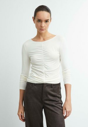 Junge Frau mit zurückgebundenem Haar trägt ein weißes gerafftes Langarmtop und dunkelbraune Hose, sie steht vor einem einfarbigen hellen Hintergrund.