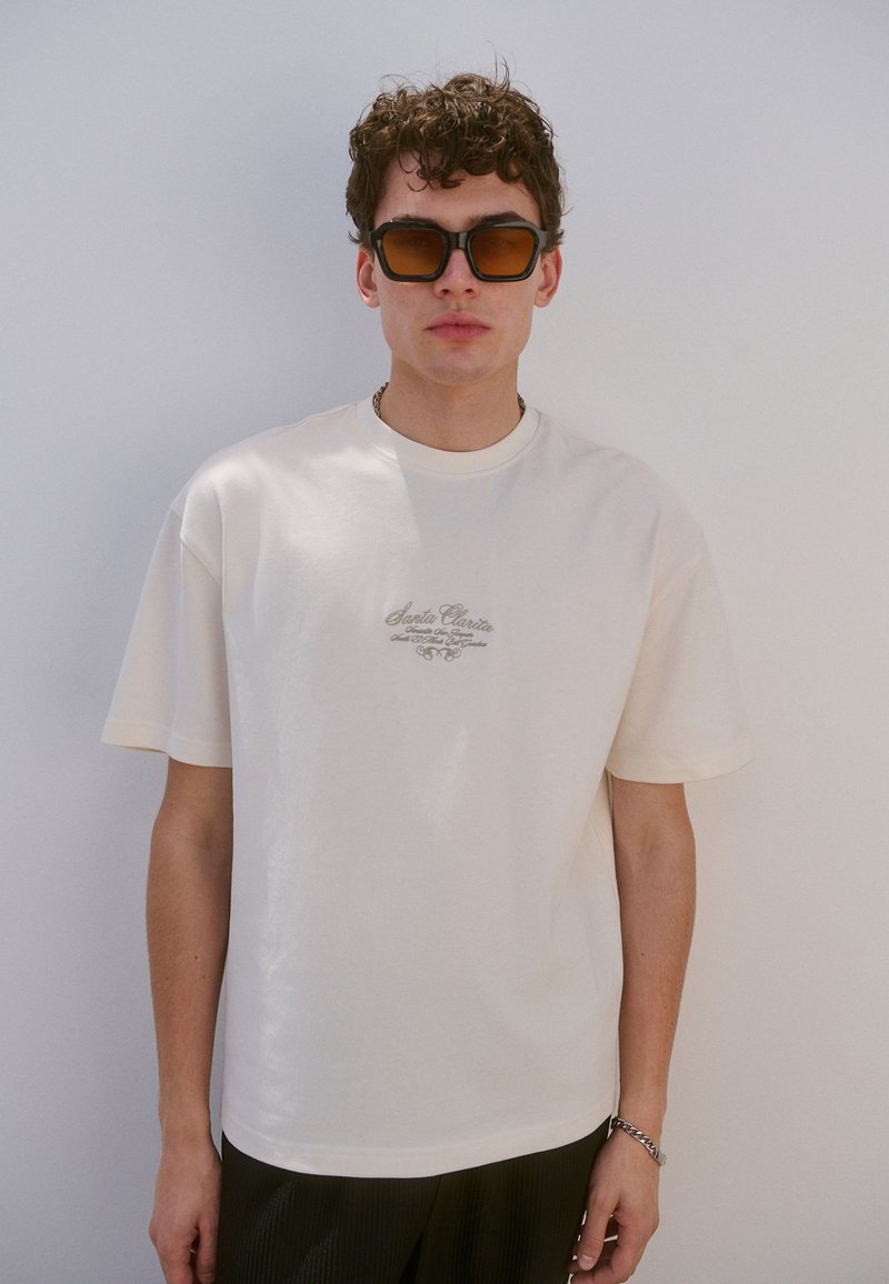 Maglietta cremisi a maniche corte in cotone, con una sottile grafica grigia con la scritta "Santa Clarita" sul petto. Tessuto liscio.