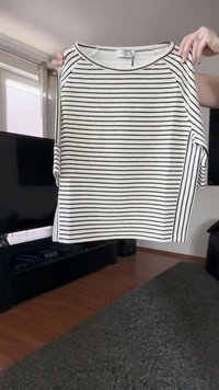 White and black horizontally striped long-sleeve shirt held up by hands in a living room with TV and carpet visible.