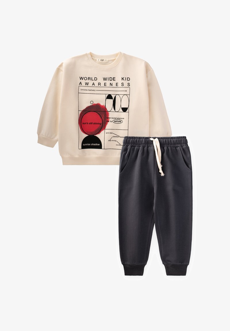 Sweatshirt beige avec impression graphique et texte, associé à un pantalon de survêtement gris foncé avec cordon de serrage, comportant des poches latérales et des poignets élastiques.