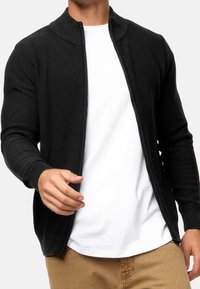 Cárdigan de punto negro con cremallera y cuello redondeado, mangas largas y puños acanalados, llevado sobre una camiseta blanca y pantalones caqui.