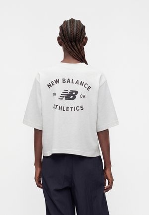 Άτομο με πλεξούδες που φοράει λευκό cropped T-shirt με το λογότυπο "New Balance 1906 Athletics" στην πλάτη και σκούρο, φαρδύ παντελόνι.