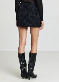 Gonna nera mini con applicazioni floreali testurizzate, vita aderente e orlo che si estende fino a metà coscia. Abbinata a stivali alti neri lucidi.
