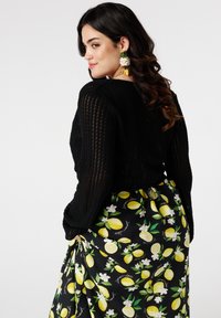 Een vrouw met lang, donker haar poseert in een zwart gebreid topje en een levendige rok met gele citroenen en witte bloemen, met speelse oorbellen.