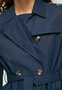 Marinblå trench coat med bälte i midjan, dubbelknäppt fram, stora bronsknappar och brett slag. Slät tyg med subtil struktur.