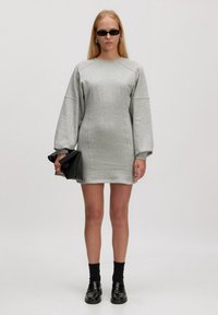 Robe-sweat grise avec de longues manches ballon, tissu texturé, coutures latérales et pochette noire. Portée avec des chaussettes noires et des mocassins.