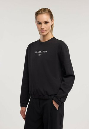 Mujer con el pelo peinado hacia atrás, que lleva una sudadera negra con el texto "TRUSSARDI 1911" y pantalones negros, de pie frente a un fondo liso.