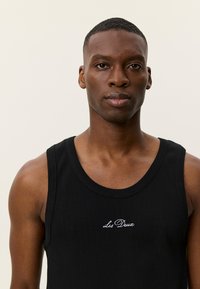 Μαύρο ριμπ τοπ με στρογγυλή λαιμόκοψη και λευκό κεντημένο λογότυπο "Les Deux" στο στήθος. Απλός σχεδιασμός, ελαφρύ ύφασμα.