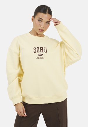 Kobieta o ciemnych włosach w kremowej bluzie z napisem "SOHO 1987 Manhattan", pozująca z jedną ręką przy czole, ubrana w brązowe spodnie.