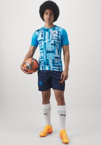 Tricou de fotbal grafic albastru și teal cu modele, șorturi bleumarin cu logo portocaliu, șosete albe și ghete de fotbal portocalii strălucitoare.