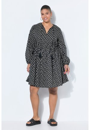 Vrouw staand en glimlachend, gekleed in een zwarte, lange jurk met patroon en franjes, zwarte slippers en grote oorbellen, tegen een witte achtergrond.