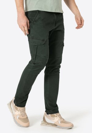 Dunkelgrüne Cargo-Hosen mit mehreren Seitentaschen, schlankem Schnitt und subtiler Textur, kombiniert mit hellbeigen Sportschuhen.