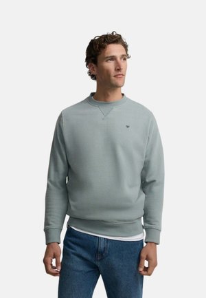 Hombre con sudadera gris claro con cuello redondo y pequeño logotipo en el pecho y vaqueros azules, de pie y mirando hacia la derecha.