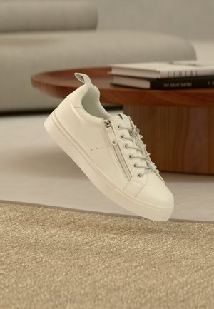 Biała niska sneaker z bocznym zamkiem błyskawicznym i sznurowadłami, umieszczona obok brązowego, okrągłego drewnianego stołu z poukładanymi książkami.