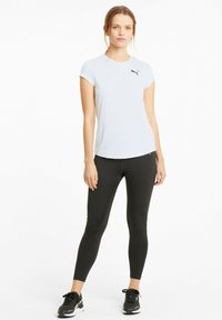 Biała koszulka sportowa z krótkim rękawem z czarnym logo Puma, w połączeniu z czarnymi legginsami i czarnymi sneakersami, na jednolitym tle.