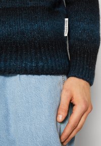 Maglione lavorato a maglia di colore blu scuro con texture a coste. Etichetta visibile sul lato. Jeans blu chiaro visibili in fondo e mano appoggiata sul tessuto.