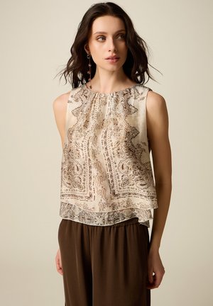 Jonge vrouw die een mouwloze beige blouse met patroon en een bruine broek draagt, kijkt naar rechts tegen een effen achtergrond.