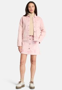 Giacca di denim rosa chiaro con due tasche frontali, abbinata a una gonna di denim rosa chiaro con bottoni frontali. Sotto, un dolcevita beige.