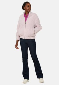 Šviesiai rožinė, pūkuota bomber striukė su užtrauktuku ir šoninėmis kišenėmis, derinama su tamsiai mėlynomis plačiomis džinsomis ir smėlio spalvos sportbačiais.